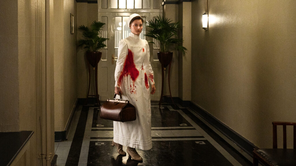 Eine Krankenschwester in weisser, blutverschmierter Kleidung und einer Arzttasche in einem Gang. 