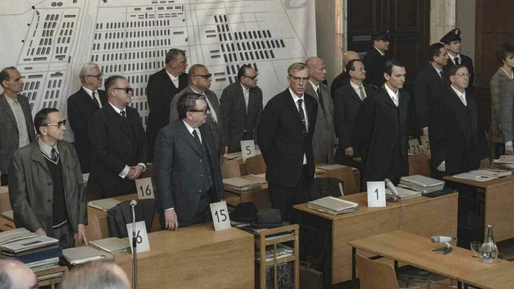 Im Gerichtssaal stehen mehrere Männer hinter Holzpulten, auf den Nummern stehen. Im Hintergrund eine schematische Karte des KZ Auschwitz.