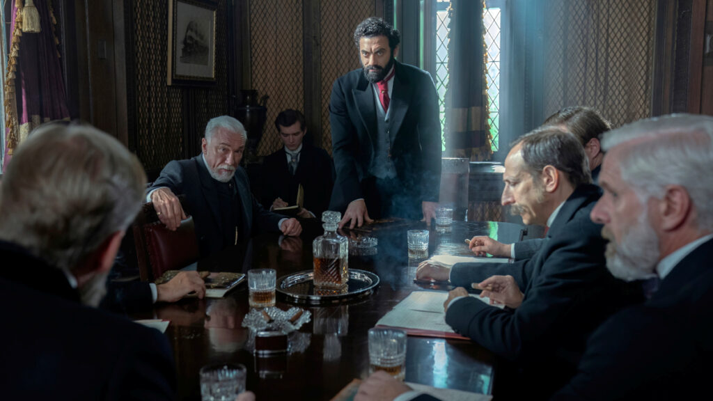 Männer in Anzügen sitzen an einem Tisch. Einer in der Mitte steht. auf dem Tisch eine Karaffe und Gläser. 