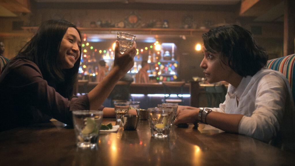 Zwei junge Frauen sitzen in einer Bar an einem Tisch, auf dem schon mehrere leere Gläser stehen. 