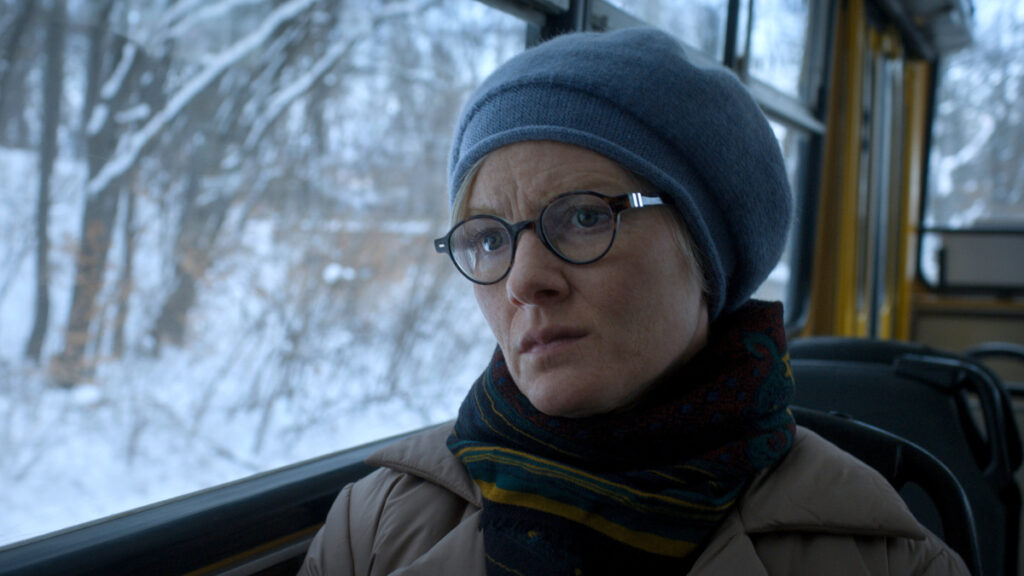 Eine Frau mit Mütze und Schal sitzt in einem Bus, der durch eine verschneite Landschaft fährt.