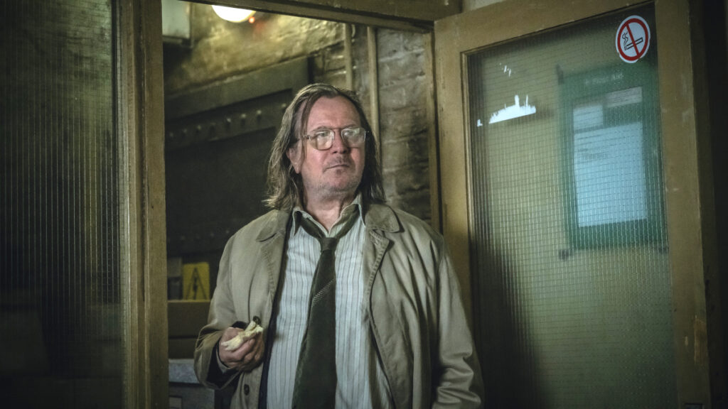 Ein Mann mit langen ungepflegten Haaren und Kleidern steht in der Tür eines heruntergekommenen Gebäudes mit einem angebissenem Sandwich in der Hand.
