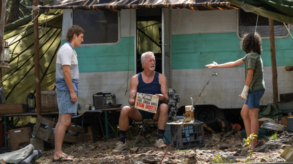 Ein älterer Mann sitzt vor einem Camper. Zwei Jugendliche reden auf ihn ein. 