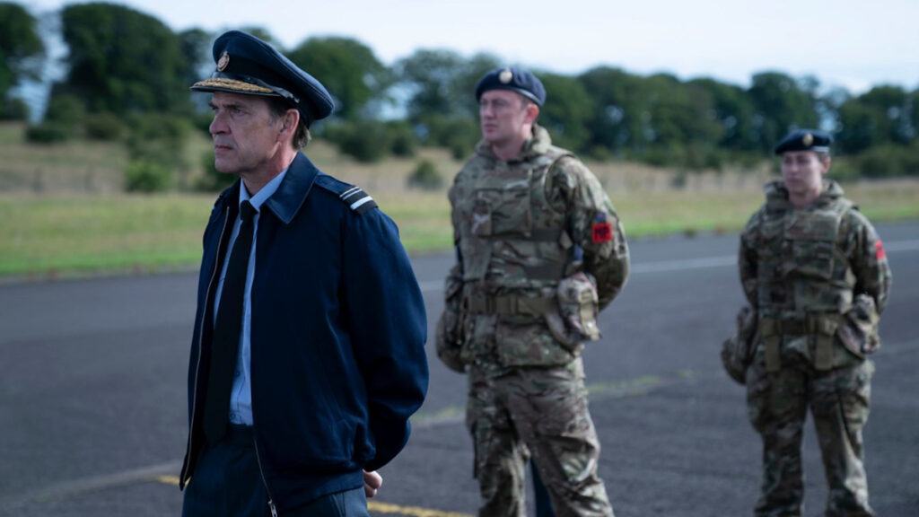 Ein Offizier in blauer Uniform steht auf einer Landebahn. Hinter ihm zwei Soldat:innen in Tarnanzügen.