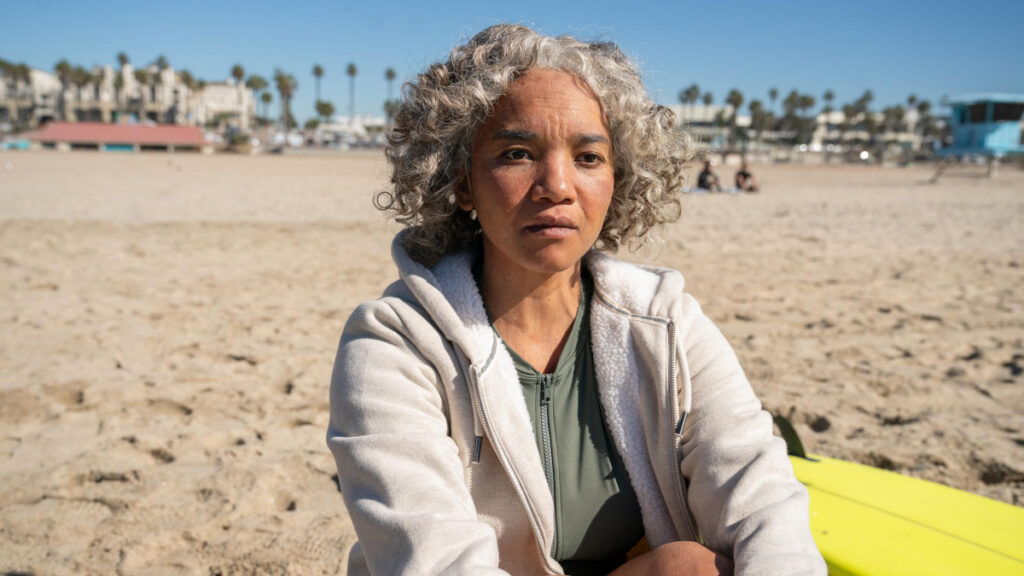 Eine weisshaarige Frau sitzt am Strand im Sand.