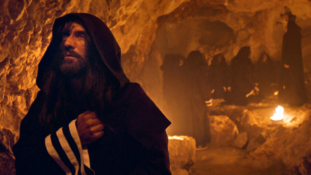 Ein Mann mit Umhang, die rechte Hand als Faust geballt auf der Brust. Dahinter in der Höhle weitere Personen in schwarzen Umhängen. 