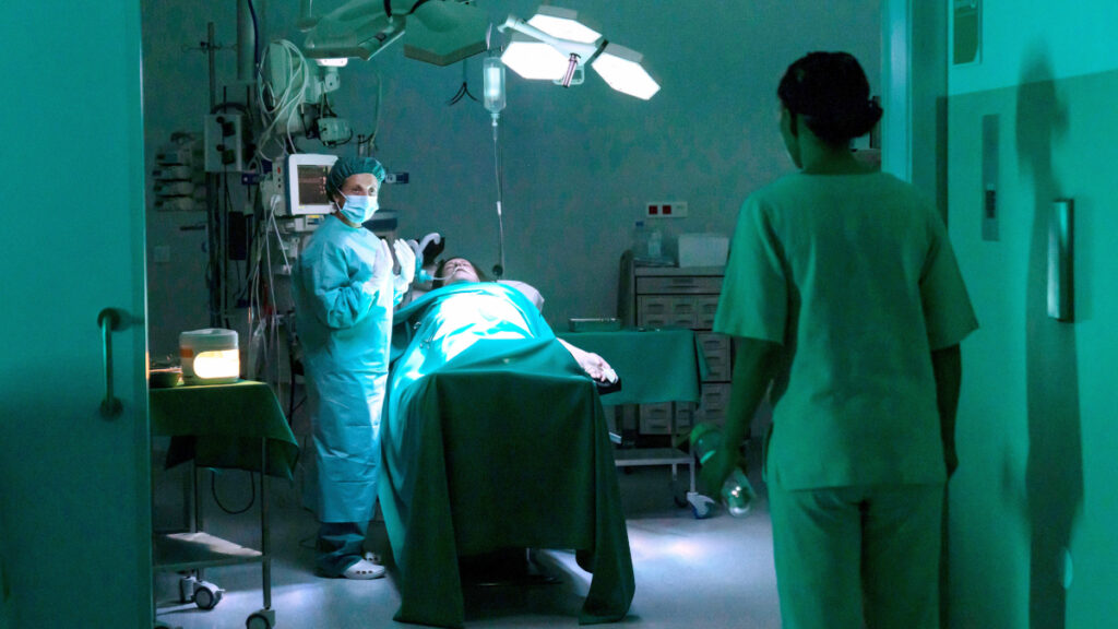 Eine Chirugin mit Mantel und Schutzmaske steht neben einem OP-Tisch, auf dem ein Patient liegt. Im Vordergrund eine Pflegerin von hinten.