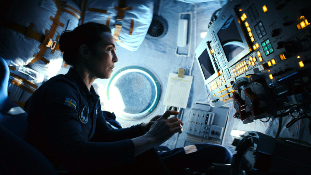Eine Frau in einer Raumkapsel vor einer Steuerungskonsole mit kleinen Bildschirmen und leuchtenden Tasten.