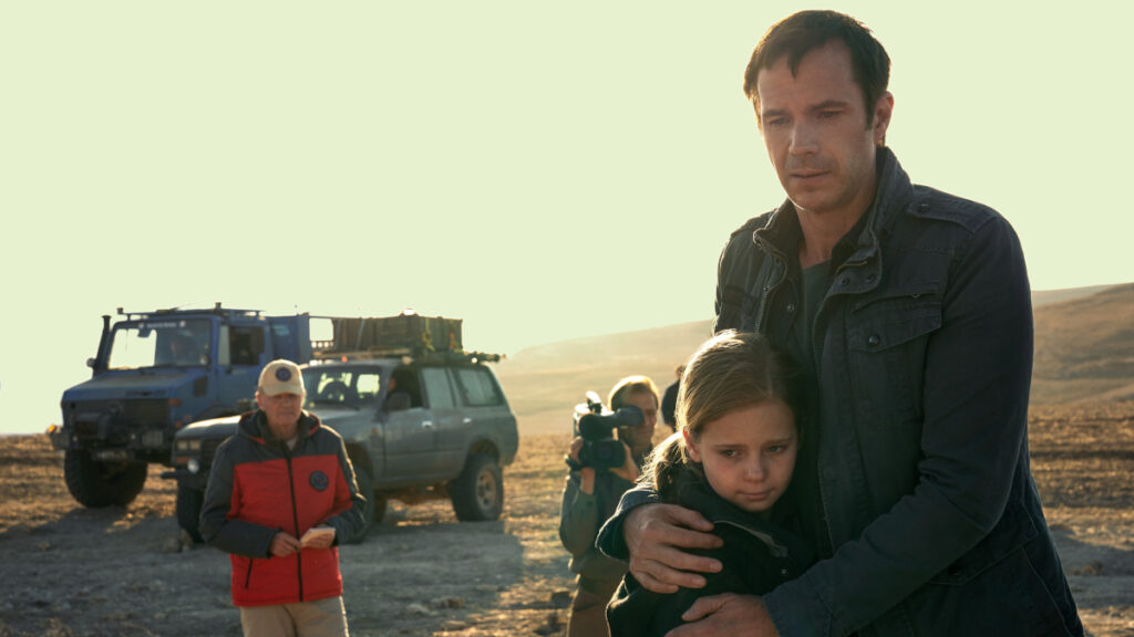 Ein Mann hält ein kleines Mädchen fest in den Armen. Sie stehen in einer kargen Landschaft, im Hintergrund zwei Fahrzeuge und ein Kameramann.
