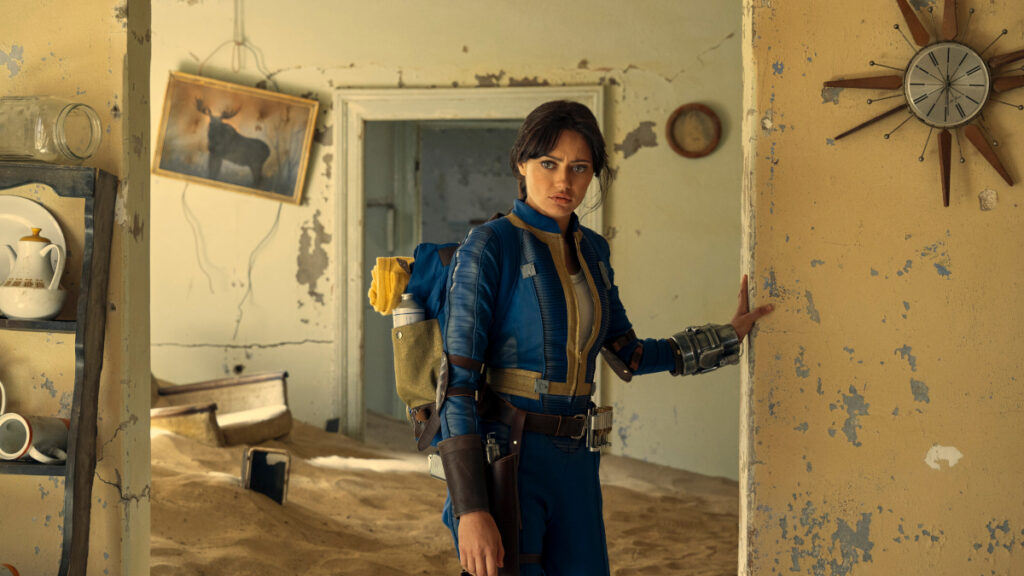 Eine junge Frau mit Rucksack und blauer Jacke steht in einem verlassenem Haus, dessen Boden völlig versandet ist.