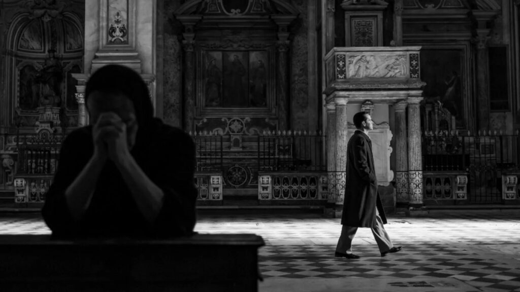 Ein Mann in einer Kirche. Im Vordergrund eine betende Frau.