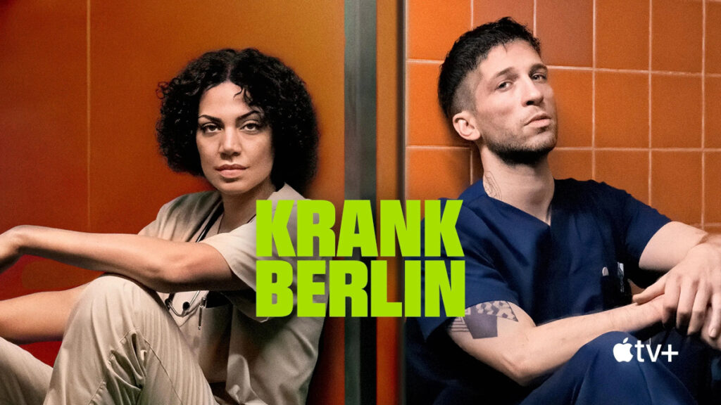 Serienpost mit Schriftzug. Eine Frau und ein Mann sitzen angelehnt an eine orange gekachelte Wand. Beide tragen Krankenhauskleidung und wirken erschöpft.