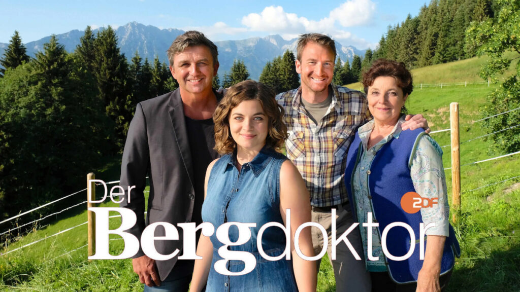 Serienposter mit Schriftzug. Zwei Männer und zwei Frauen vor einer bergigen, grünen Landschaft.