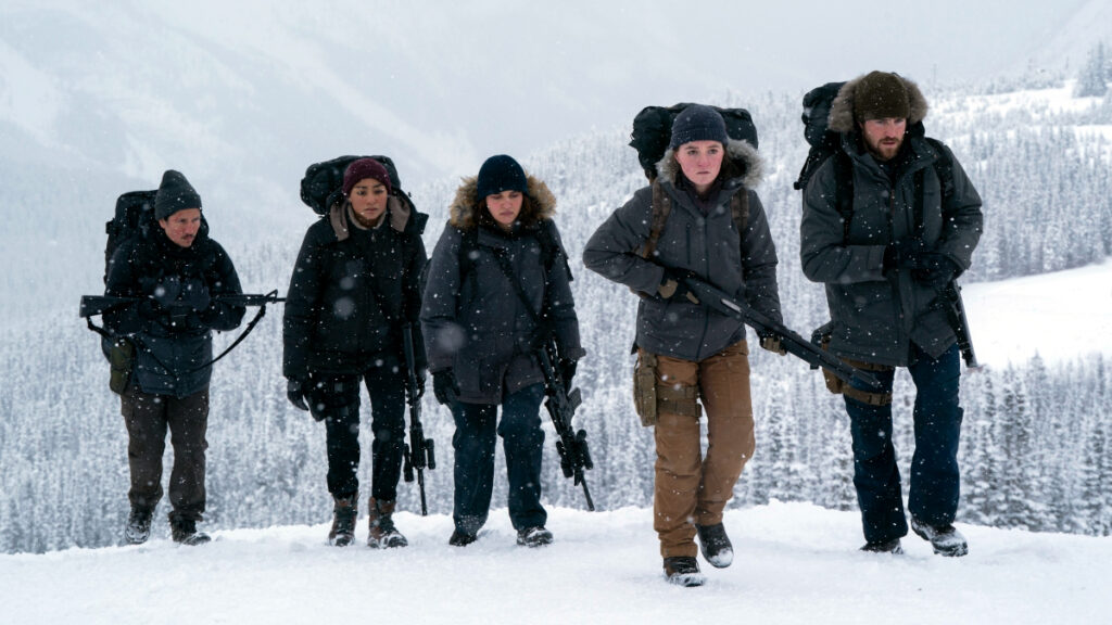 Fünf Menschen mit Rucksäcken und Waffen in einer verschneiten Landschaft.