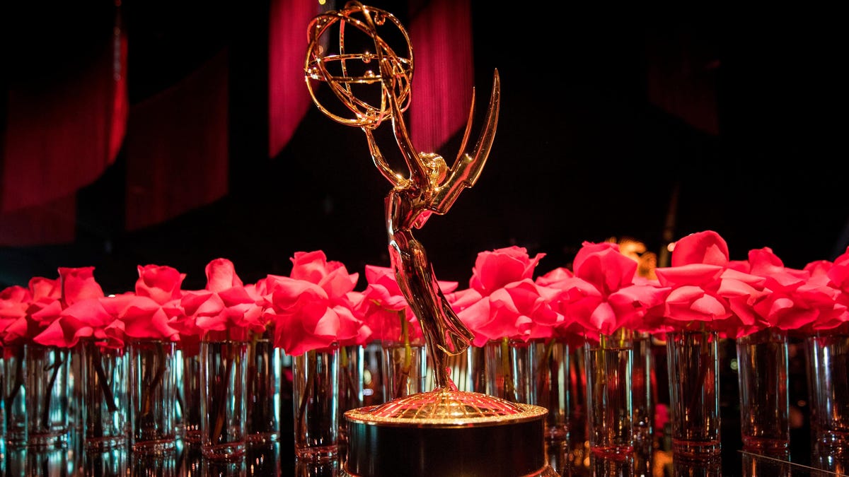 Eine goldene Trophäe steht im Vordergrund, umgeben von leuchtend roten Rosen in Vasen. Der Hintergrund ist dunkel.