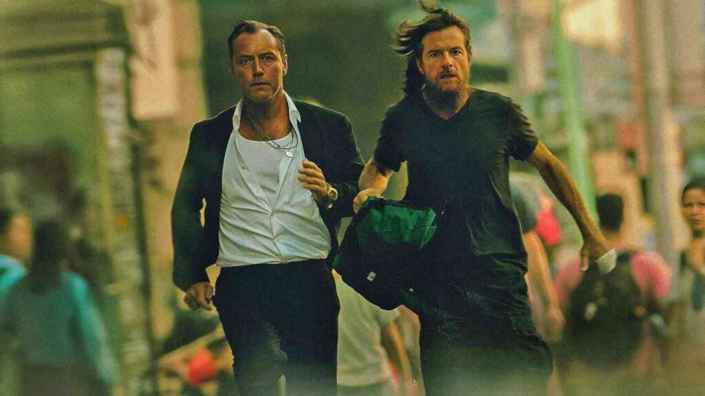Zwei Männer rennen durch eine belebte Strasse. Der linke trägt einen Anzug mit offenem Hemd, der rechte ein schwarzes Shirt und hält eine Tasche. Die Umgebung ist urban und verschwommen, mit Passanten im Hintergrund. Die Szene wirkt angespannt und dynamisch.