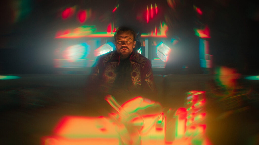 Ein Mann sitzt in einem düsteren Raum. Die Szene wirkt durch bunte Lichter psychedelisch verzerrt. Er trägt einen auffälligen roten Anzug mit geometrischem Muster. Im Hintergrund sind unscharfe Lichter und Bildschirme zu sehen, die eine mystische Atmosphäre erzeugen.
