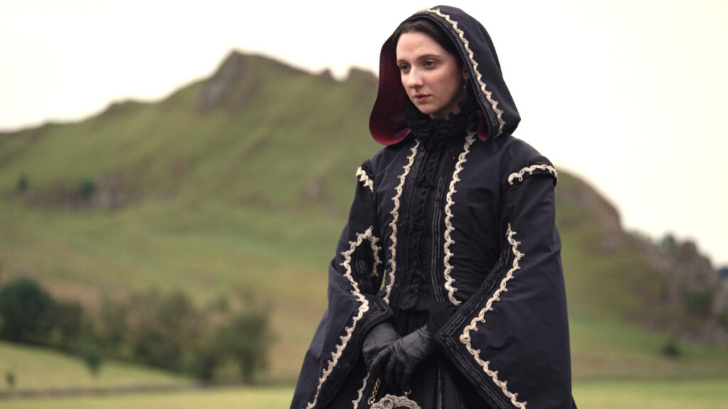 Eine Frau in historischer Kleidung mit besticktem Mantel und Kapuze steht vor einer grünen, hügeligen Landschaft. Sie wirkt nachdenklich und blickt zur Seite. Im Hintergrund sind unscharf Bäume und Hügel erkennbar. Die Szene vermittelt eine ruhige, melancholische Stimmung.