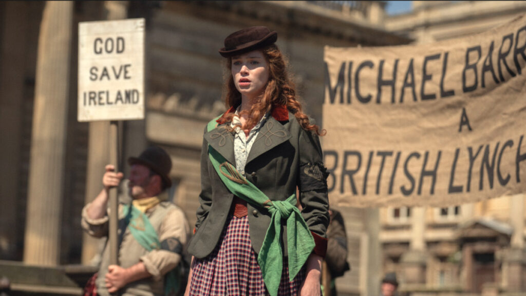 Eine Frau in historischer Kleidung steht bei einer Protestszene. Sie trägt einen Hut und ein grünes Halstuch. Im Hintergrund sind Demonstranten mit Bannern zu sehen, auf denen englische Schriftzüge wie «God Save Ireland» und «Michael Barr A British Lynch» zu lesen sind.