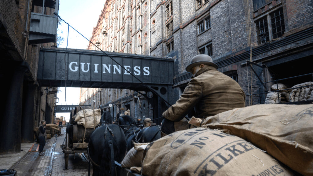 Eine historische Strassenszene mit Pferdekutschen, beladen mit Säcken, die durch eine enge Gasse unter einer Brücke mit der Aufschrift «GUINNESS» fahren. Das Kopfsteinpflaster und die alten Backsteingebäude schaffen eine authentische, zeitgenössische Atmosphäre.