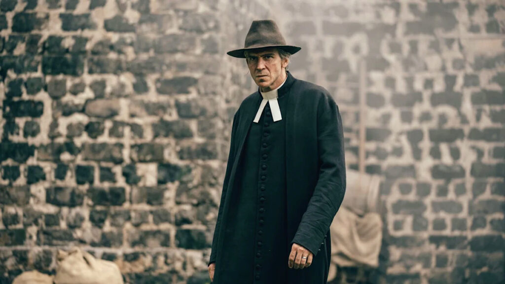 Ein Mann in Priesterkleidung steht vor einer alten Steinmauer. Er trägt einen braunen Hut und schaut ernst in die Kamera. Der Hintergrund zeigt unscharf weitere Gegenstände und trägt zur düsteren Atmosphäre der Szene bei.