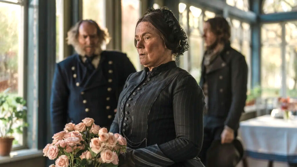Eine Frau in viktorianischer Kleidung hält einen Strauss rosa Rosen. Im Hintergrund stehen zwei Männer in einem hellen Raum mit grossen Fenstern. Die Szene wirkt historisch und dramatisch.