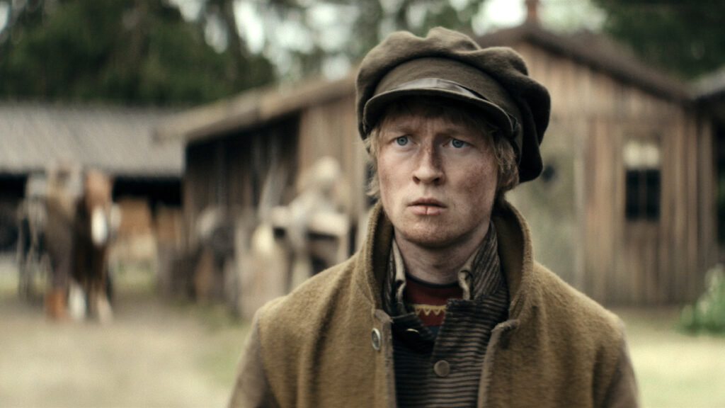 Ein junger Mann mit ernstem Ausdruck steht im Freien vor einem unscharfen Hintergrund aus Holzgebäuden. Er trägt eine braune Schiebermütze und einen Mantel. Die Szenerie wirkt ländlich und historisch.