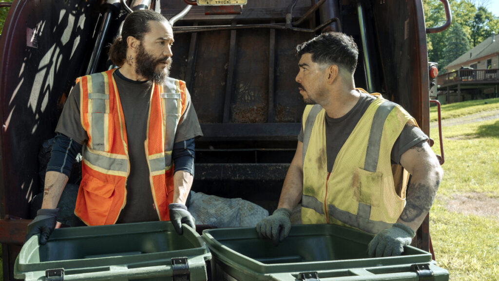 Zwei Männer in Arbeitskleidung und Sicherheitswesten stehen vor einem Müllwagen. Sie halten Mülltonnen und sind in ein Gespräch vertieft. Im Hintergrund ist eine grüne Landschaft zu sehen.