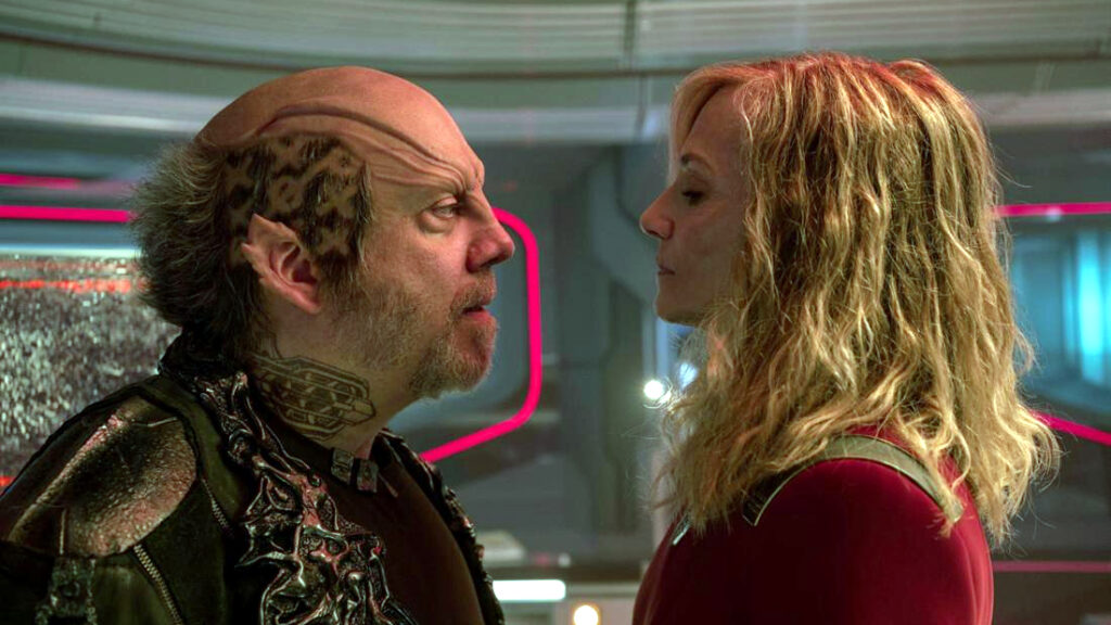 Eine Person mit alienartigem Aussehen und eine Frau in einem futuristischen Raum stehen sich intensiv gegenüber. Der Hintergrund zeigt leuchtende Linien und technologische Elemente. Die Szene wirkt konfrontativ und spannend.
