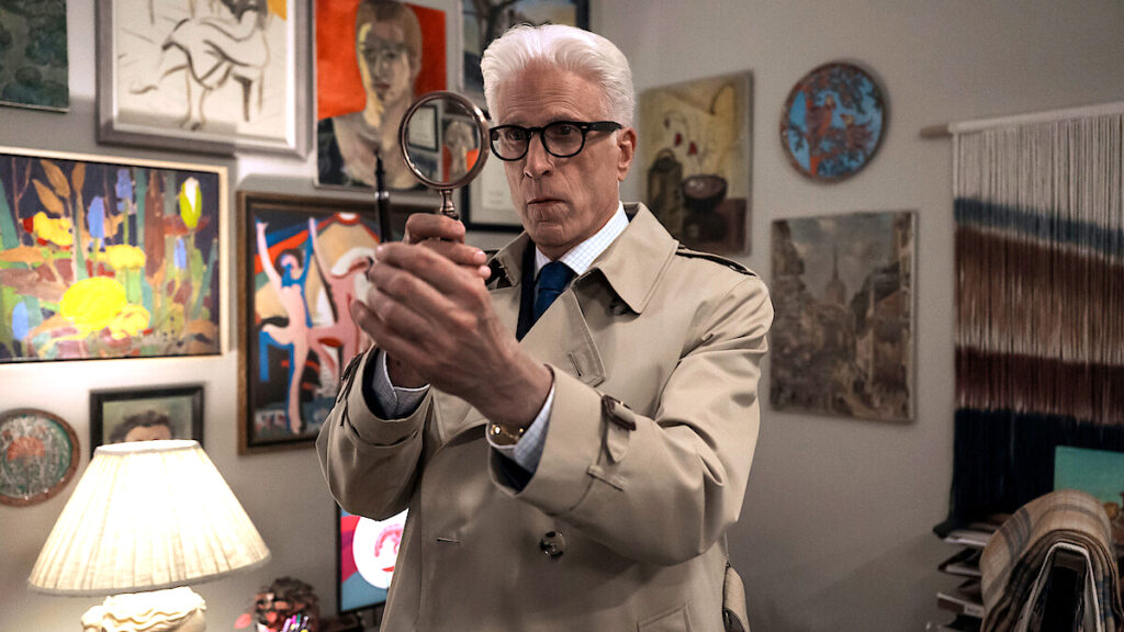 Ein Mann in Trenchcoat steht in einem Raum voller bunter Kunstwerke und hält eine Lupe vor sein Gesicht. Eine Tischlampe beleuchtet den Raum, der eine gemütliche, kreative Atmosphäre ausstrahlt. Die Wände sind mit Gemälden und Wandbehängen dekoriert.