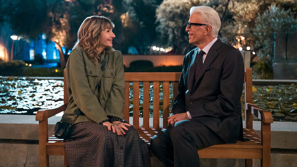 Ein Mann und eine Frau sitzen auf einer Parkbank vor einem Teich in nächtlicher Umgebung und unterhalten sich lächelnd. Die Frau trägt eine Jacke und einen langen Rock, der Mann einen Anzug. Im Hintergrund sind Bäume und Lichter zu sehen.