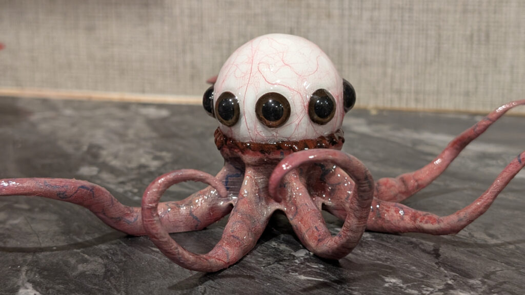 Oktopus-ähnliches Wesen mit kugelförmigem, weissem Kopf und mehreren schwarzen Augen. Die Tentakel sind rosa und mit blauen Adern durchzogen. Die Figur steht auf einer marmorierten Fläche vor einer strukturierten, grauen Wand.