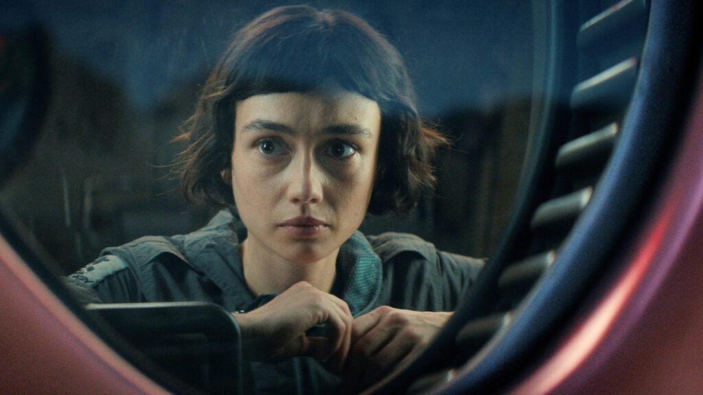 Eine Person mit kurzem, dunklem Haar schaut ernst durch ein rundes Fenster. Sie trägt eine dunkle Jacke. Die Umgebung wirkt futuristisch oder technisch. Das Bild vermittelt eine nachdenkliche oder spannende Stimmung.