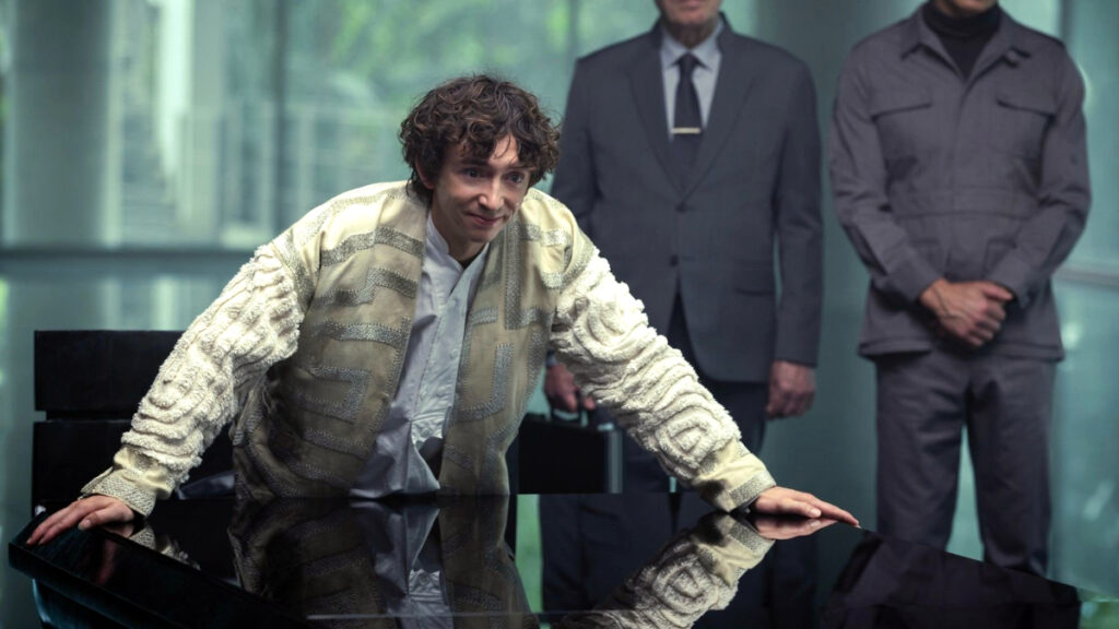 Eine Person mit lockigem Haar lehnt sich lächelnd über einen dunklen Tisch. Sie trägt eine gemusterte helle Jacke und ein weisses Hemd. Im Hintergrund stehen zwei weitere Personen in Anzügen, die keine Emotionen zeigen, in einem modernen Raum mit grossen Glasflächen.