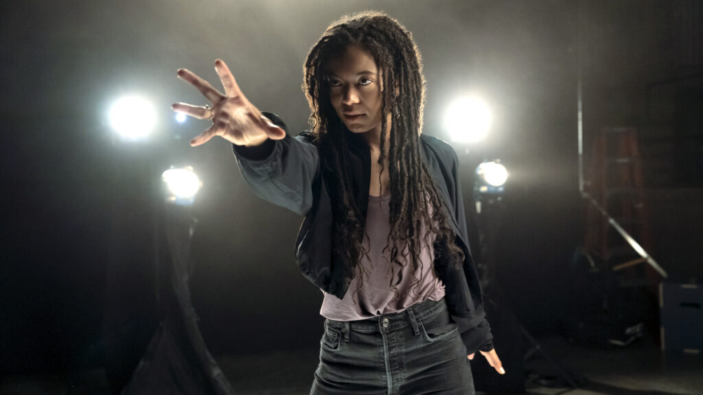 Eine Person mit langen, lockigen Haaren und lässiger Kleidung streckt dynamisch die Hand nach vorne. Im Hintergrund sind drei helle Lichter zu sehen, die eine dramatische Atmosphäre schaffen.
