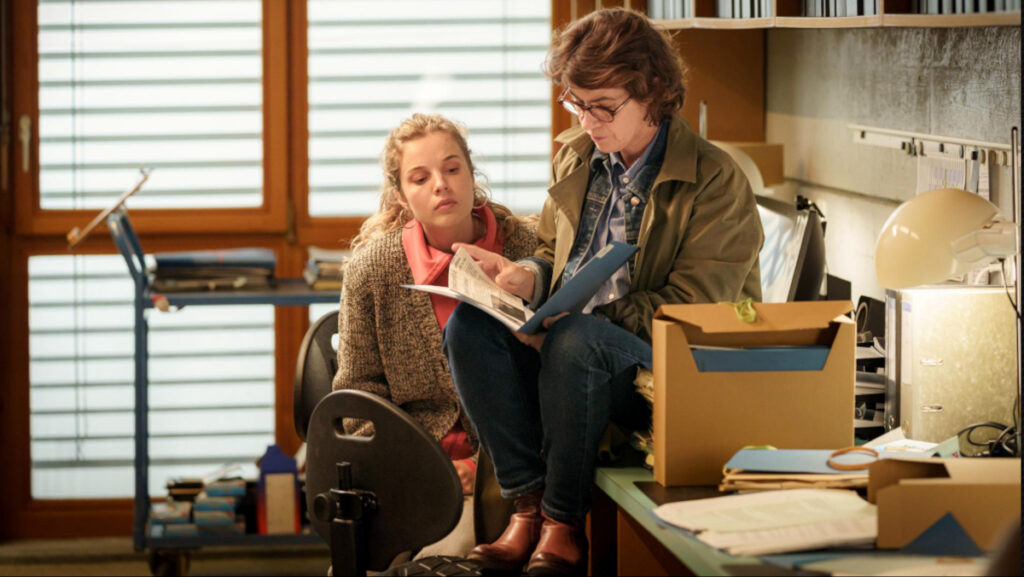 Zwei Personen befinden sich in einem Büro. Eine sitzt auf einem Bürostuhl, die andere auf dem Tisch. Beide betrachten gemeinsam Akten. Um sie herum sind Schachteln, Ordner und eine Lampe zu sehen. Der Raum wird von aussen durch ein Fenster mit Jalousien beleuchtet.