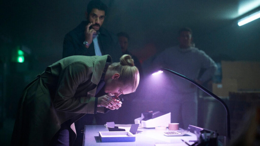 Eine Person im Mantel untersucht konzentriert mit einer Lupe ein Objekt unter einer Lampe in einem abgedunkelten Raum. Zwei weitere Personen sind im Hintergrund unscharf zu erkennen. Der Tisch ist mit technischen Geräten und Papieren bedeckt. Die Szene wirkt geheimnisvoll und intensiv.