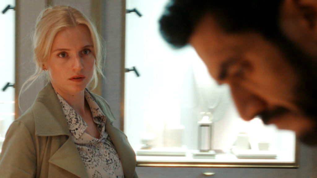 Eine Frau mit blondem Haar steht in einem hell beleuchteten Raum und schaut ernst. Im Vordergrund ist verschwommen ein Mann zu sehen. Im Hintergrund sind Vitrinen mit Schmuckstücken ausgestellt. Die Szene wirkt angespannt.