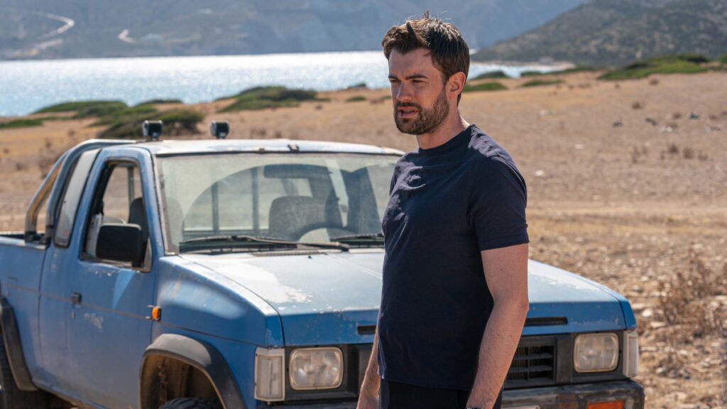 Ein Mann steht vor einem blauen Pickup-Truck in einer trockenen, leicht hügeligen Landschaft mit Blick auf ein Gewässer. Im Hintergrund sind Hügel und Büsche zu sehen. Der Himmel ist klar und sonnig.
