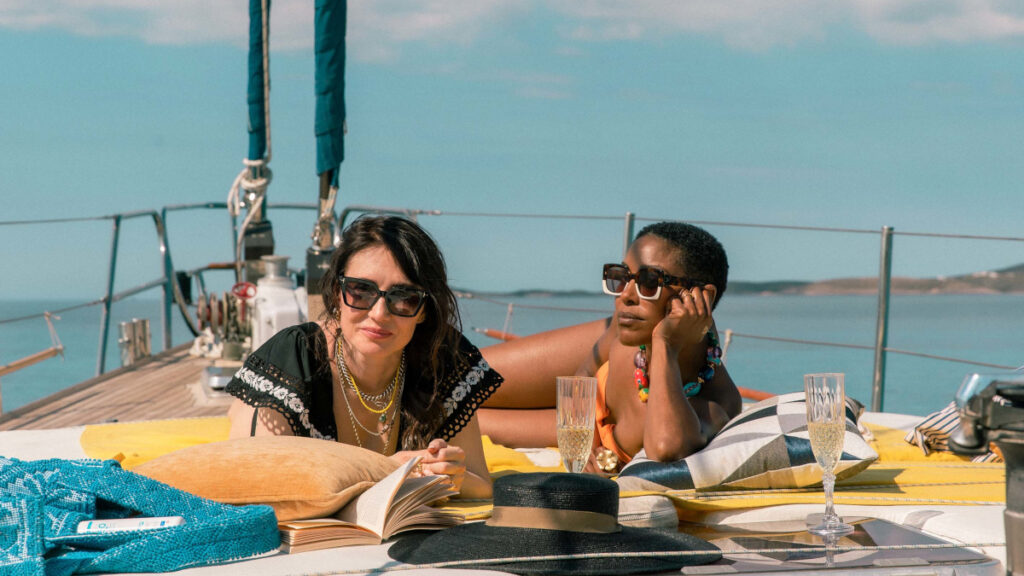 Zwei Frauen entspannen auf einem Segelboot. Sie liegen auf einem Sonnendeck mit Kissen, lesen ein Buch und geniessen ein Getränk. Der Himmel ist blau, und im Hintergrund ist das Meer sichtbar. Beide tragen Sonnenbrillen und schauen entspannt.