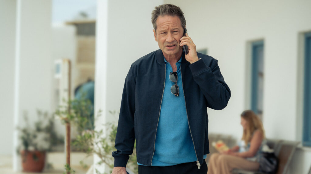 Ein Mann in einem blauen Hemd und dunkler Jacke telefoniert draussen vor einem weissen Gebäude. Sonnenbrille hängt am Hemd. Im Hintergrund sitzt eine andere Person auf einer Bank und liest. Pflanzen dekorieren den Bereich.
