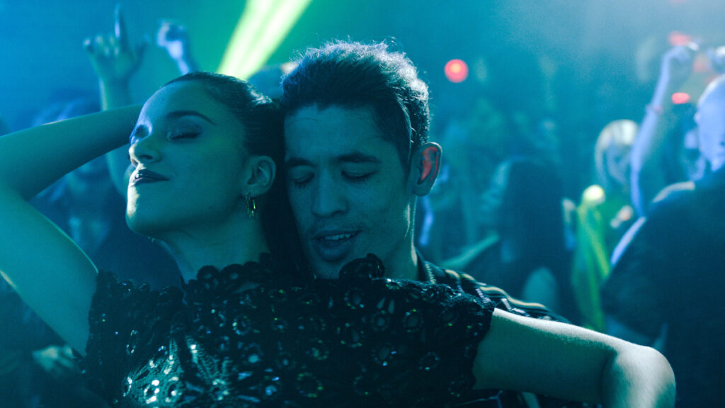 Eine Frau und ein Mann tanzen eng umarmt in einem Club. Die Frau hat die Augen geschlossen, wirkt entspannt und trägt ein schwarzes Kleid mit Pailletten. Der Mann hat ebenfalls die Augen geschlossen. Im Hintergrund sind weitere tanzende Personen und bunte Lichter zu sehen. Stimmung: lebhaft und ausgelassen.