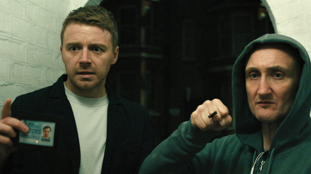 Zwei Männer stehen in einem schmalen Durchgang. Der linke Mann hält eine Ausweiskarte und blickt ernst. Der rechte Mann trägt einen Kapuzenpullover und zeigt eine Geste mit der Hand. Die Szene wirkt angespannt und findet in einer dunklen Umgebung statt.