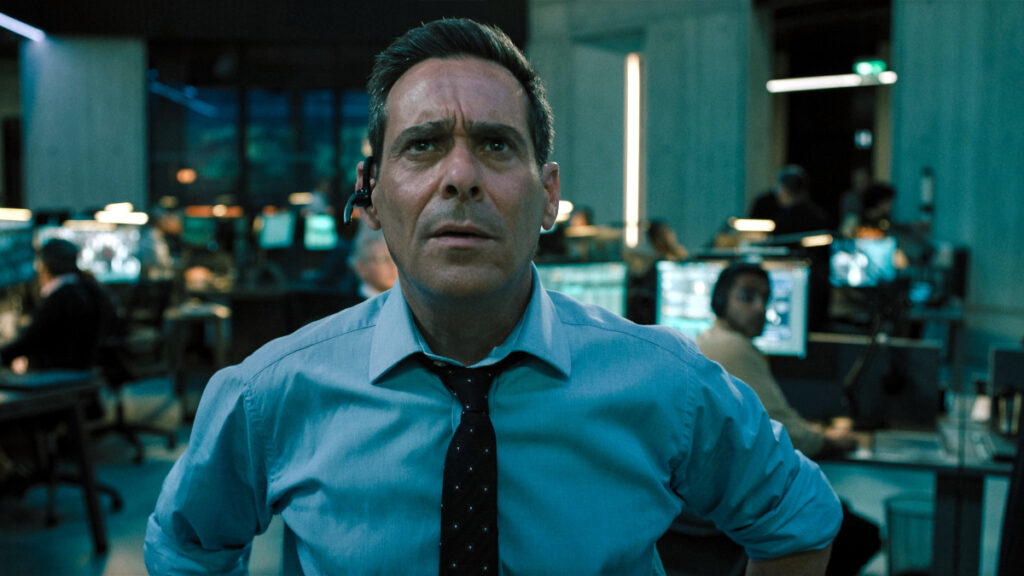 Ein Mann in einem Büro steht im Vordergrund und blickt ernst in die Ferne. Er trägt ein Headset und ein hellblaues Hemd mit Krawatte. Im Hintergrund sind mehrere Arbeitsplätze mit Menschen und Monitoren zu sehen. Sie befinden sich in einem grossen, modernen Kontrollraum.