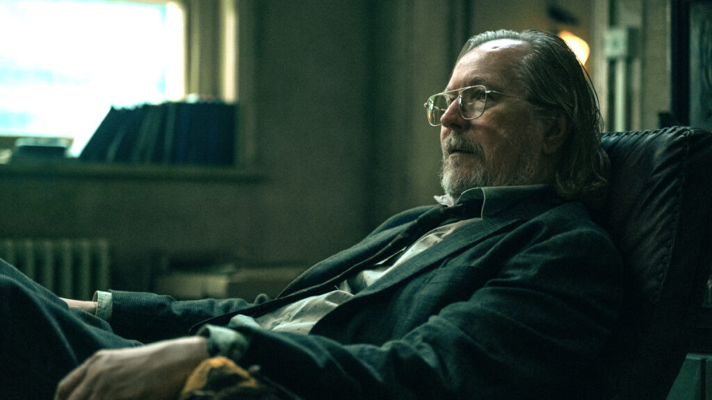 Ein Mann mit Brille und mittellangem, grauem Haar sitzt nachdenklich in einem Sessel in einem schummrig beleuchteten Raum. Im Hintergrund sind unscharfe Bücherregale und ein Fenster sichtbar. Die Stimmung ist ruhig und nachdenklich.