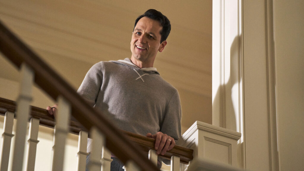 Ein Mann in einem grauen Kapuzenpullover steht lächelnd auf einer Treppe und schaut nach unten. Die Umgebung scheint ein helles, elegantes Gebäude zu sein, da man ein weiss gestrichenes Geländer und Wände sieht. 