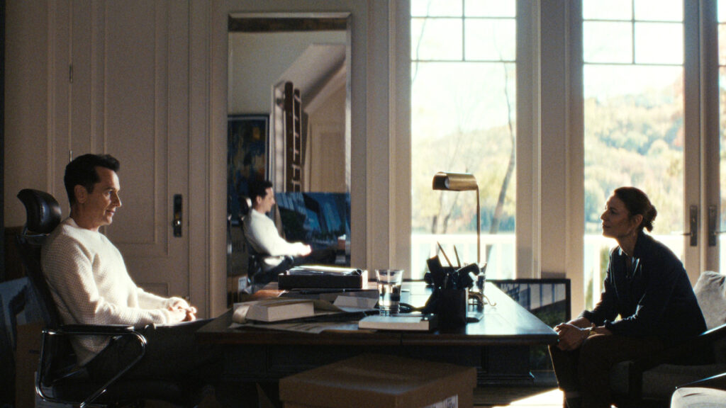 Zwei Personen sitzen sich an einem Schreibtisch gegenüber, in einem hellen Raum mit Blick auf mit Bäumen bewachsene Hügel. Im Vordergrund liegt Büromaterial, darunter Bücher und eine Lampe. Ein Spiegel an der Wand reflektiert eine der sitzenden Personen.