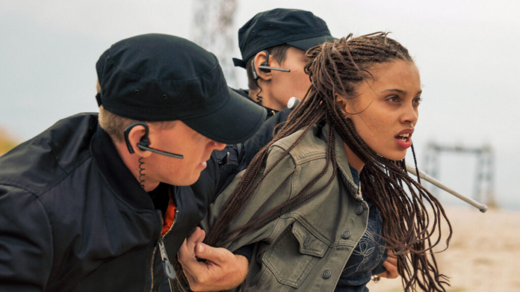 Zwei Personen in schwarzen Mützen und Jacken mit Headsets halten eine Frau mit geflochtenen Haaren fest. Die Frau wirkt angespannt und ist im Profil abgebildet. Der Hintergrund zeigt einen unscharfen Strand oder eine offene Fläche.