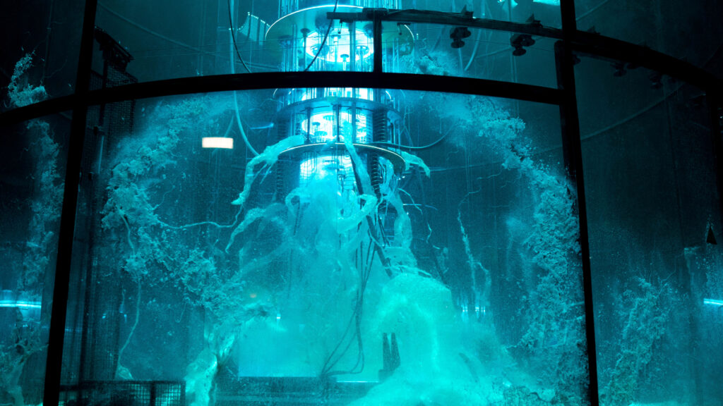 Das Bild zeigt ein futuristisches Labor mit einem grossen zylindrischen Tank, gefüllt mit blau leuchtender Flüssigkeit. Drähte und leuchtende Elemente erstrecken sich im Inneren. Die Szene vermittelt eine Atmosphäre von Wissenschaft und Technologie, möglicherweise für Experimente oder Forschung.