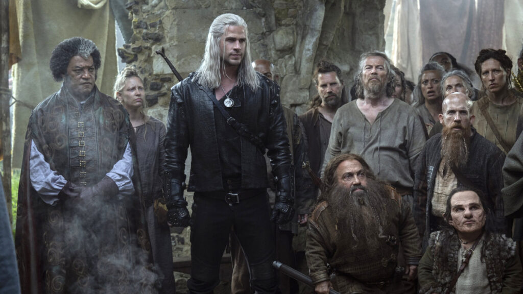 Eine Gruppe von Menschen in mittelalterlichen Gewändern steht in einer steinernen Umgebung. Im Zentrum steht eine Person mit langen, weissen Haaren in schwarzer Lederkleidung. Umgeben von verschiedenen Charakteren, einige blicken in die Ferne, andere direkt in die Kamera.